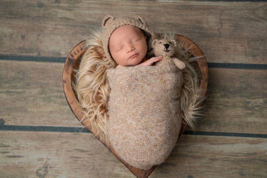 Newborn baby in a bear bonnet, tucked into a heart-shaped bowl with a knit teddy bear