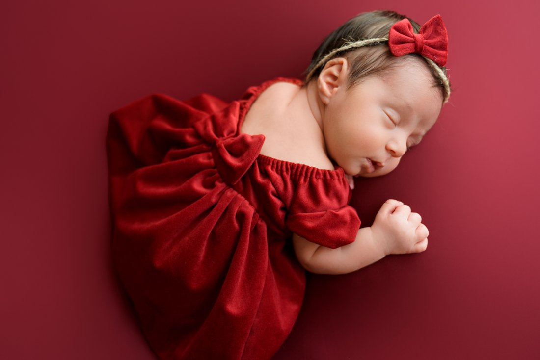 Newborn baby in a red velvet dress and bow headband, sleeping on a deep red backdrop